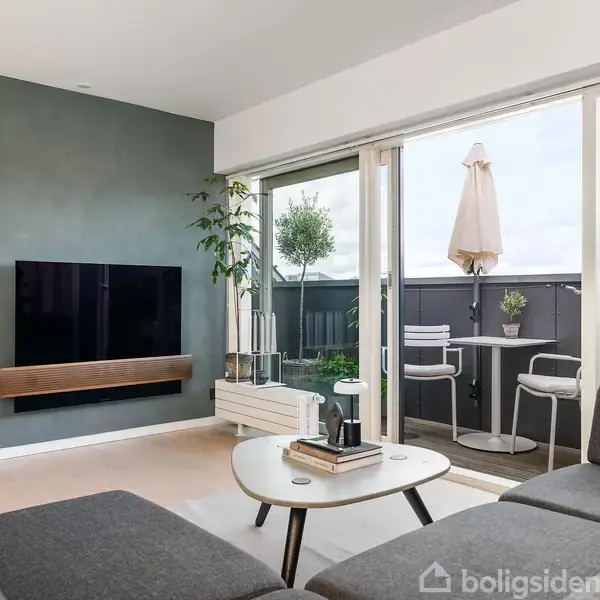 Stue med grå sofaer og et rundt sofabord, hvorpå der står en kande. Tv monteret på en grøn væg. Store vinduer fører ud til en altan med borde og stole.