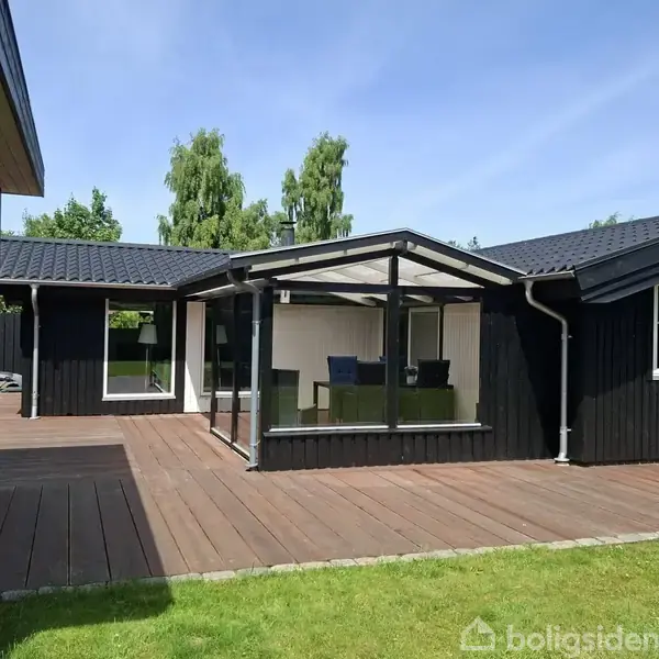 Sortmalet hus med terrasse af træ, omgivende græsplæne og træer i baggrunden. Gennemsigtigt udestuerum med stole og bord indeni, beliggende i et roligt, grønt område.