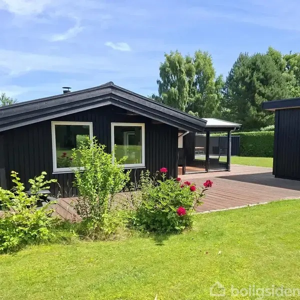 Sort træhus ligger stille på en grøn græsplæne med blomsterbede i forgrunden. Til venstre er der buske, og i baggrunden ses frodige træer under en blå himmel.
