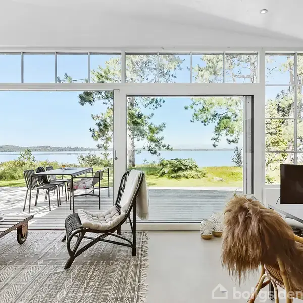 Stol står i lys stue med store vinduer. Udsigt til terrassebord og stole udenfor, omgivet af græs og træer samt udsigt til vand og himmel.