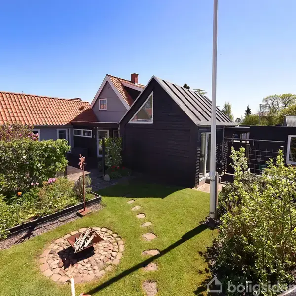 Et lille hus med spidstag står i en solrig have med en brolagt sti og en bålplads. Grønt græs og buske omkranser området.