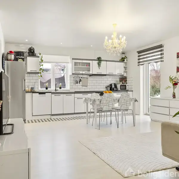 Et stilfuldt køkken med hvidt interiør har et spisebord med gennemsigtige stole. En lysekrone hænger fra loftet, og store vinduer giver udsigt til en have.