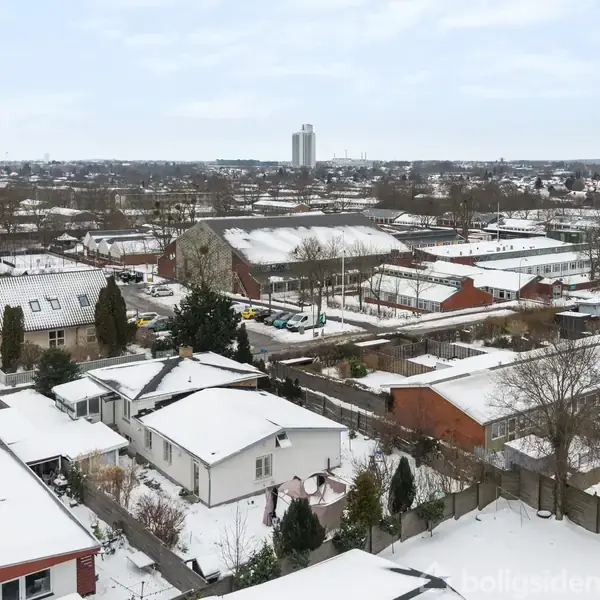 Hustage er dækket af sne i en bydel med mange enfamiliehuse. Sneen ligger også på vejene og haverne, og i baggrunden ses en høj bygning mod en klar himmel.