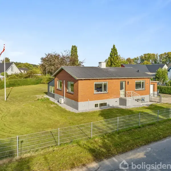 En rødstensvilla står stille på en velplejet græsplæne i et villakvarter med flere træer omkring og et hegn langs vejen.