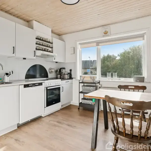 Et lyst køkken med hvide skabe og træbord, hvor stole står omkring. En stor vindue giver udsigt til træer. På bordet er der en kaffemaskine og andre køkkenredskaber.