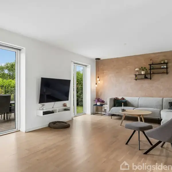 Stue med hjørnesofa og sofabord, vægmonteret fladskærm på lys væg. Glaspaneler leder ud til terrasse med havemøbler. Hylder med dekorationer hænger på væggen ved sofaen.