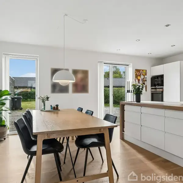 Træbord omgivet af sorte stole står i et lyst køkken. Hvide skabe og en køkkenø ses ved siden af store vinduer med udsigt til en have. En lampe hænger over bordet.