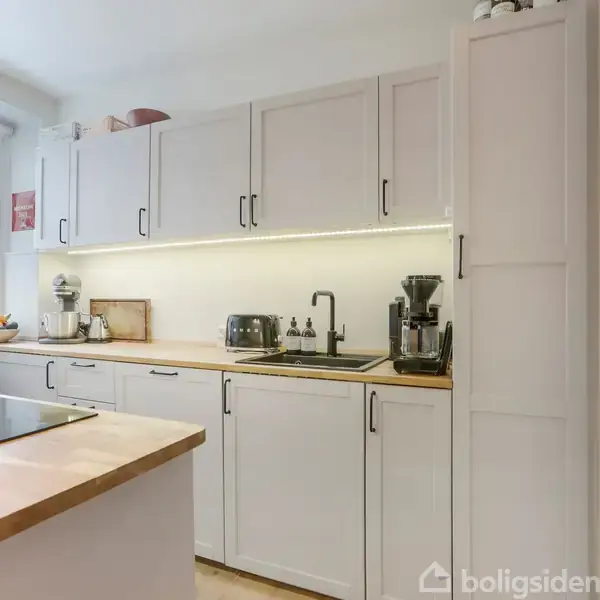 Et lyst køkken med lyse træskabe og bordplade. Der står en brødrister og kaffemaskine på bordet. Et stort vindue til venstre giver udsyn til natur udenfor.