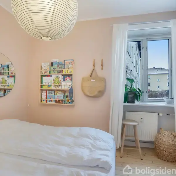 En seng med hvidt sengetøj står i et soveværelse. Ved siden af er en væg med en rund spejl, en hylde med bøger og en taske hængende. En stol og plante er ved vinduet. En rund lampe hænger fra loftet.