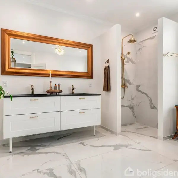 Badeværelse med dobbeltvask på hvid skabsenhed, stor spejlramme i træ, bruseområde med marmorlignende fliser og separat badekar. Rummet er lyst og elegant med moderne indretning.