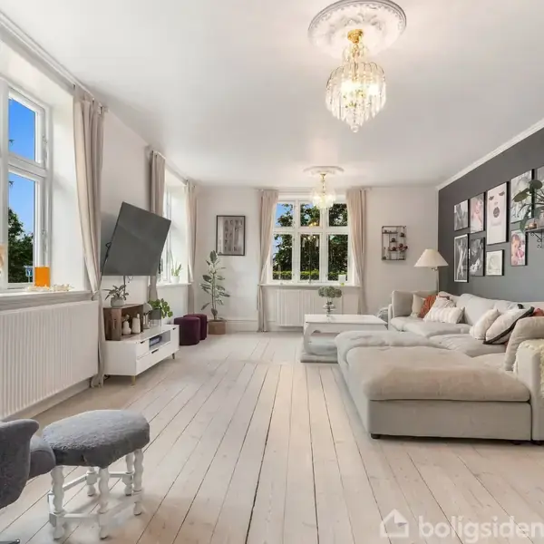 Stue med stor beige sofa, fladskærms-TV på væggen og chandelier i loftet. Væggene er hvide, med et mørk accentvæg, dekoreret med billeder. Store vinduer med udsigt til træer.