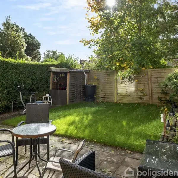 Haveområde med terrassebord og stole står på flisebelagt område. Græsplæne omgivet af hæk og træer. Træskur og kompostbeholder i baggrunden. Sollys skinner gennem trækronerne.