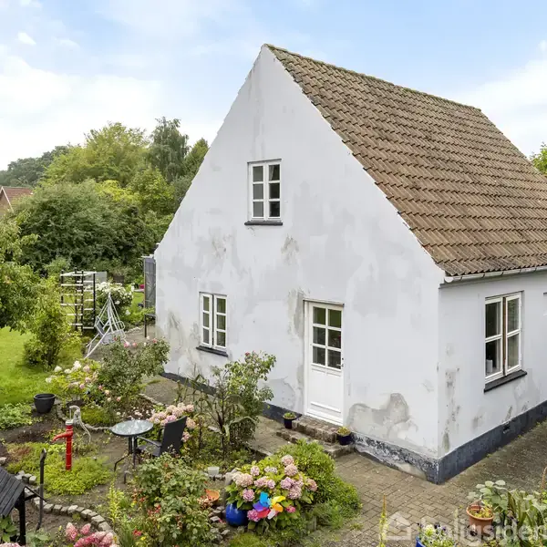 Hvidmalet hus med skrå tagsten, står i en frodig have med blomsterbede og træer, en stensti fører til indgangsdøren.