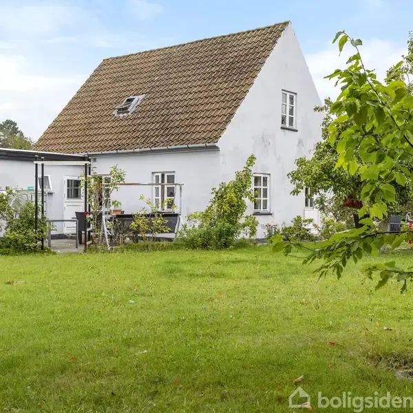 Hvidt hus med rødt tag står i grøn have. Træer og buske omgiver huset, som har en terrasse med bænk. Himmel er blå med få skyer.