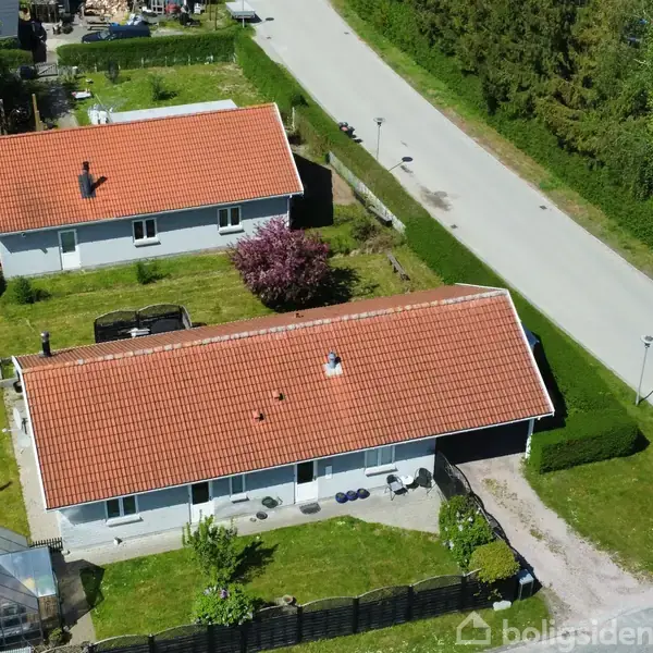 To ens-etagers huse med røde tage ligger ved en vej med træer og buske omkring. Der er en have med drivhus og en blomstrende busk mellem husene.