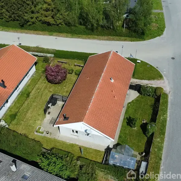 To huse med røde tage ligger tæt ved en vej i et boligområde. Haverne er grønne med buske og træer, indrammet af hække, og et drivhus ses ved det ene hus.