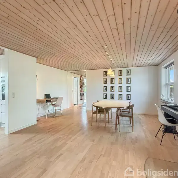 Stuen har et spisebord med stole, en pejs i hjørnet og et skrivebord ved vinduet. Trægulve og træloft skaber en varm atmosfære. Væggen har indrammede billeder.