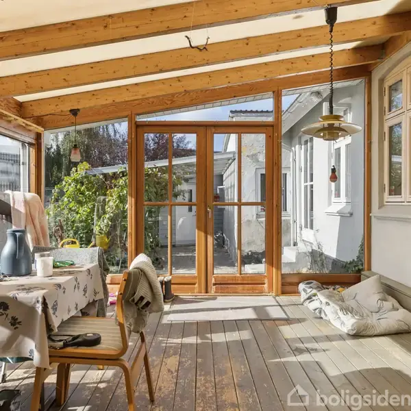 En veranda med træmøbler og et bord dækket med en dug. Sollys strømmer ind gennem glasvægge, og planter ses udenfor. Der hænger en lampe fra loftet.