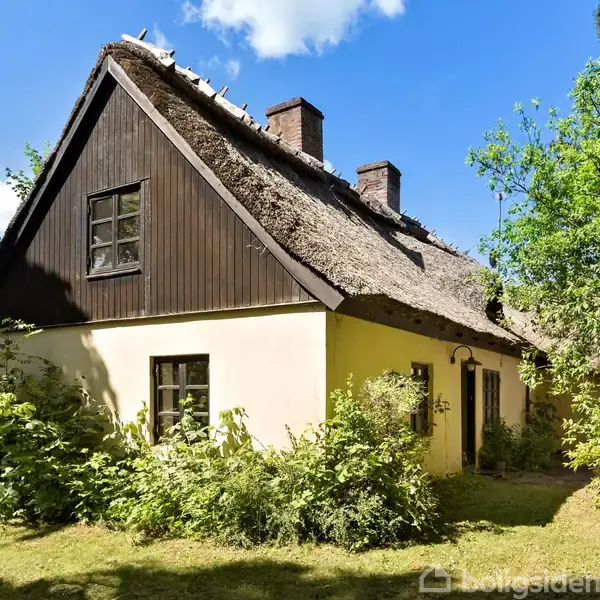 Et stråtækt hus står stille omgivet af grønne buske og træer. Den solbeskinnede himmel giver et fredfyldt landskab, hvor naturen omfavner bygningen.
