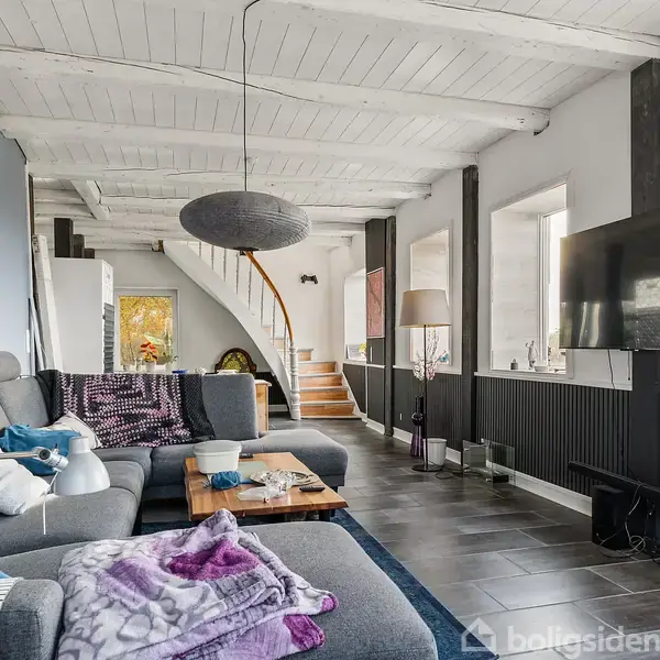 En grå sofa med puder står i en stue med hvidt træloft. En fladskærm er monteret på væggen, og trappen i baggrunden fører til en øvre etage.