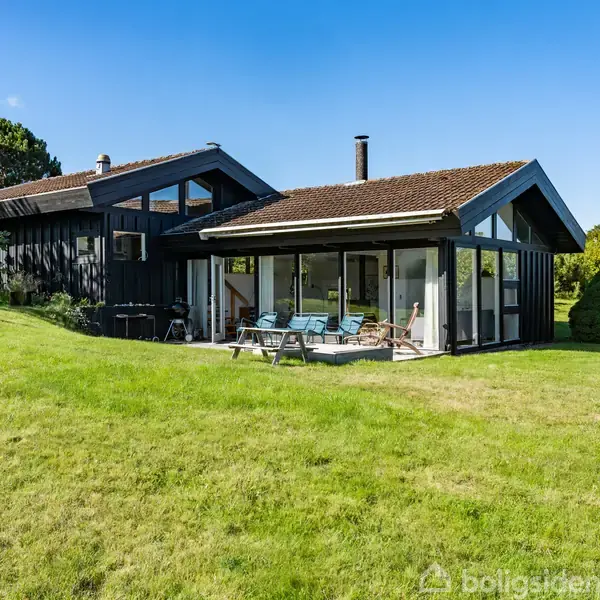 Et moderne sortmalet sommerhus med store vinduer står stille. Det er omgivet af en velplejet græsplæne og omgivet af træer og buske under en klar blå himmel.