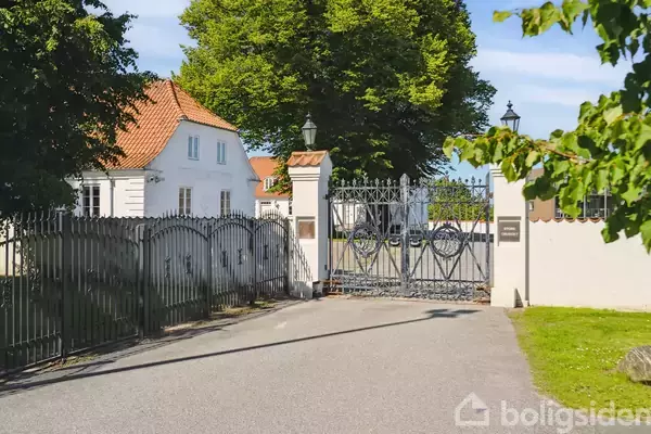 En smedejernslåge står åben på en grusvej, omgivet af en hvid bygning med rødt tegltag og grønne træer.