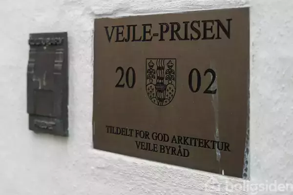 Et skilt fastgjort på en væg. Det viser teksten "VEJLE-PRISEN 2002" samt et byvåben. Nedenfor står: "TILDELT FOR GOD ARKITEKTUR VEJLE BYRÅD".