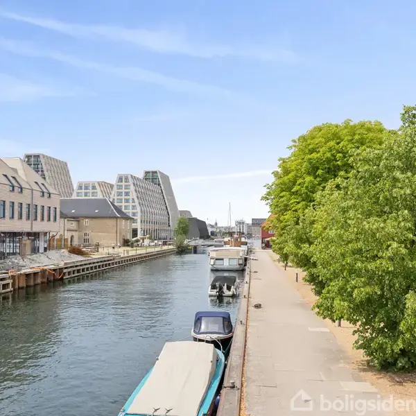 Kanal løber mellem moderne bygninger på venstre side og en promenade med træer på højre. Både ligger fortøjet ved kanten af vandet under en klar, blå himmel.
