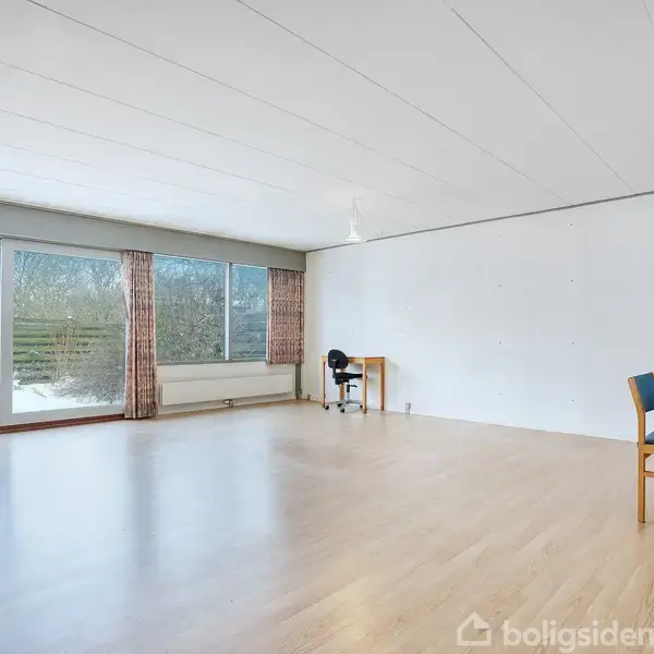 En stor, tom stue med lyst trægulv og store vinduer mod en snedækket have, indeholder et skrivebord med stol og en kommode med to stole i hjørnet.