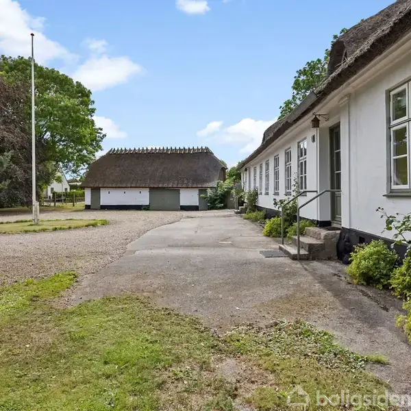 Hvid bygning med stråtag står langs en gårdsplads med grus. Der er en anden mindre bygning med stråtag i baggrunden, omgivet af træer og grønt.