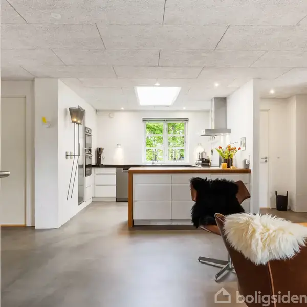 Et lyst køkken med en central ø, omgivet af moderne apparater. På en brun lænestol ligger et hvidt fåreskind. Store vinduer giver udsigt til grønne træer udenfor.