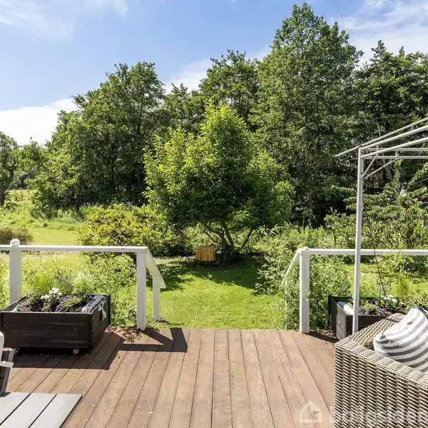 Terrasse med havemøbler og blomsterkasser, omgivet af en grøn have med træer. En klar blå himmel ses i baggrunden, der skaber en fredfyldt atmosfære.