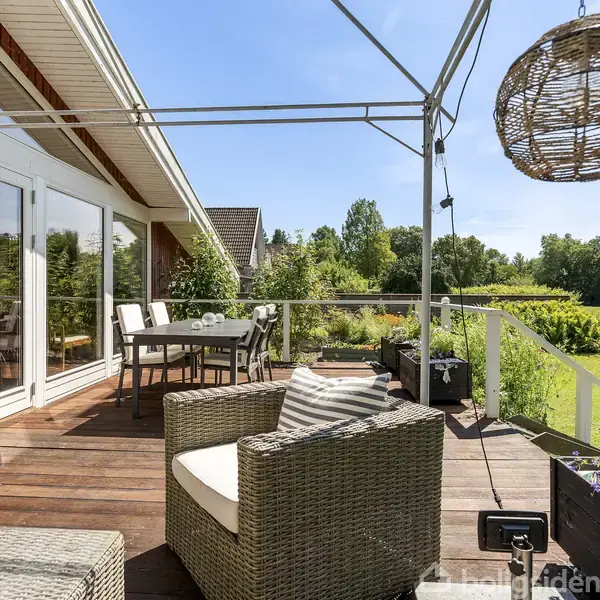 Terrassemøbler står på en træterrasse med udsigt over en frodig have. En hængestol er monteret i en metalramme, mens lyse hynder pryder stolene. Fjern grøn baggrund.