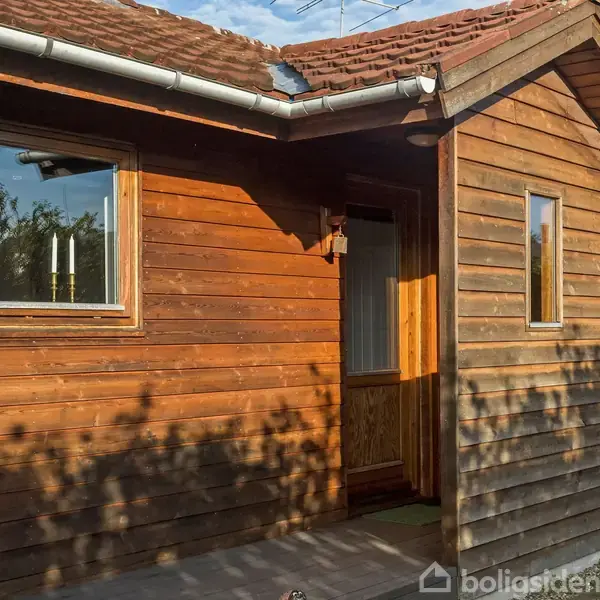 Et træhus med rødt tag og vinduer oplyst af sollys. Omgivet af grønne træer og buske i en landlig have.