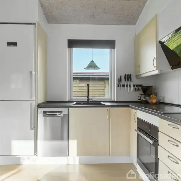 Et lyst og moderne køkken med køleskab, opvaskemaskine og ovn, placeret under hvide og træfarvede skabe. En stor vask er under et vindue, der giver udsyn til udendørsområdet.