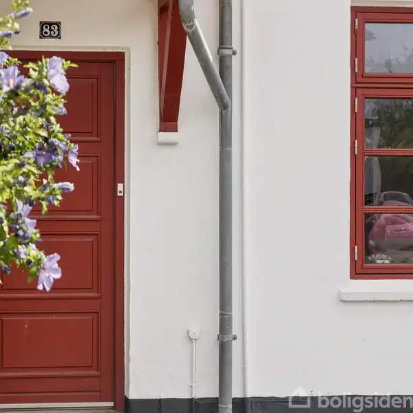 Rød hoveddør med nummer 83 ved siden af et vindue. En grøn busk med lilla blomster vokser til venstre. Dør og vindue er på en hvid husfacade.