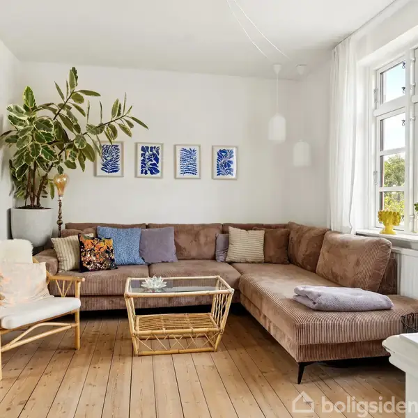 Brun hjørnesofa med puder står i en stue. Ved siden af står en lænestol og et sofabord. Vægkunst og grøn plante pryder væggen. Store vinduer lukker lys ind.