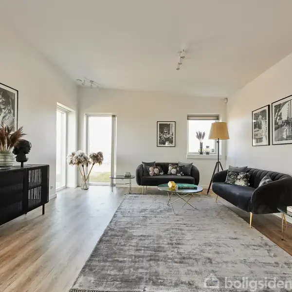 To sorte sofaer står i en lys stue med trægulv, en stor grå tæppe og minimalistisk indretning. Vægkunst og tørrede planter pryder rummet.