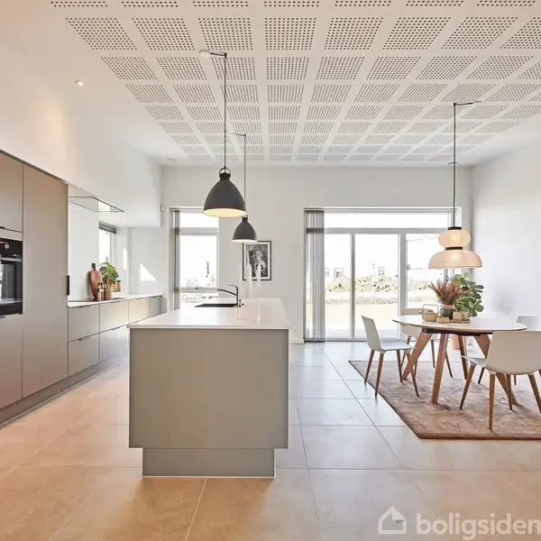Et moderne køkken-alrum har en stor køkkenø med pendellamper ovenover og et spisebord med fire stole på et tæppe. Rummet er lyst og minimalistisk indrettet.