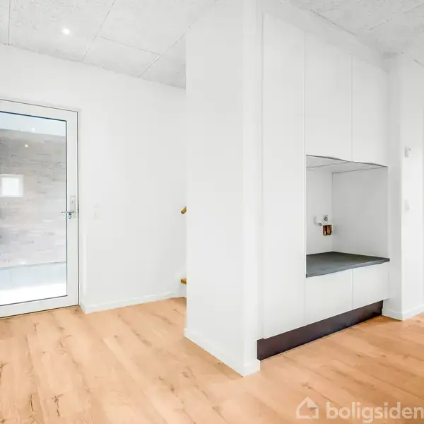 Dør til badeværelse set fra gang. Lys træparketgulv dominerer rummet, med et indbygget skab i højre side. Véggene er hvide, og loftet er pletbelyst.