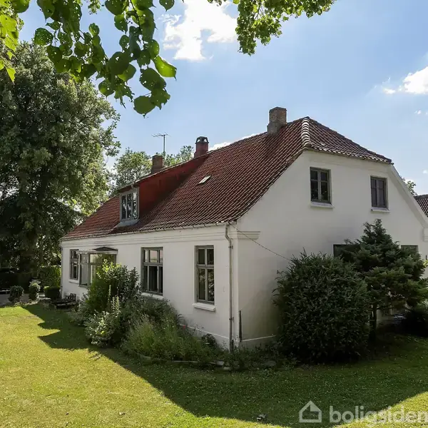 Et hvidt hus med rødt tegltag står i en grøn have med træer og buske. Skyfri blå himmel i baggrunden.