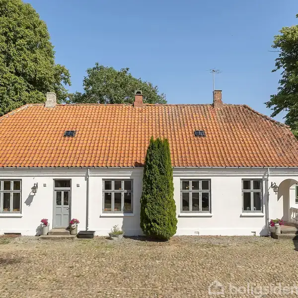 Hvidt hus med rødt tegltag står stille på en gårdsplads omgivet af grønne træer under en klar blå himmel. Flere vinduer og en hoveddør ses på forsiden.