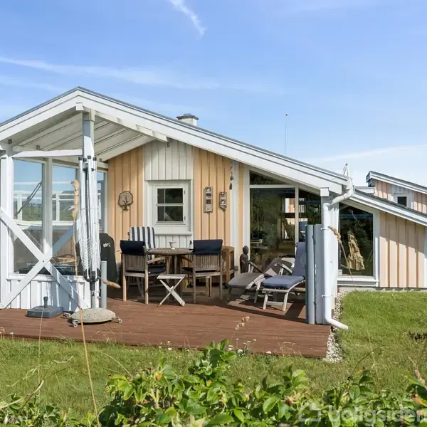Sommerhus med træterrasse, udstyret med havemøbler, står i en solrig, grøn have. Vinduer og døre giver lys til det lyse træinteriør. Blå himmel og en hængekøje ses i baggrunden.