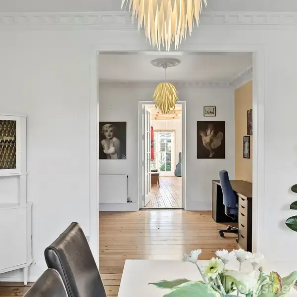 En spisebord med sorte stole står i et lyst rum; et dekorativt loftlys hænger over det. I baggrunden ses et kontorområde med træbord og kunst på væggene.