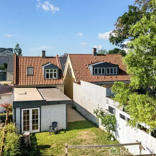 To huse med røde tegltage står side om side, med en lille have imellem. En græsplæne og et hvidt stakit omgiver husene, og et træ kaster skygger.