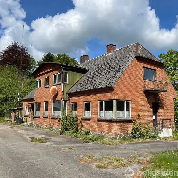 Et rødt murstenshus med flere skæve vinkler står stille i en frodig, skovklædt omgivelse under en blå himmel med hvide skyer.