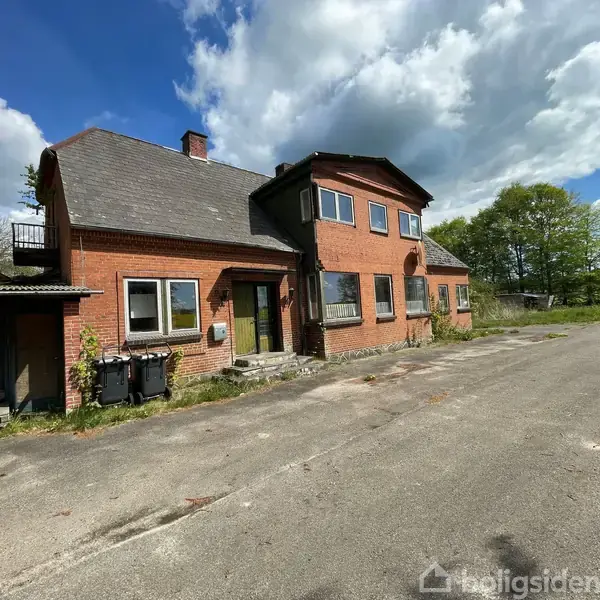 En to-etagers rød murstensbygning står ved en asfalteret vej omgivet af grønne træer under en delvist skyet himmel. Ingen synlig tekst.