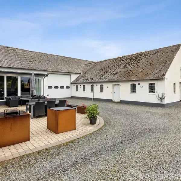 Et hvidt landhus med grå tage står stille i en stor gårdsplads. Gårdspladsen er omgivet af brostensbelægning og haveanlæg.