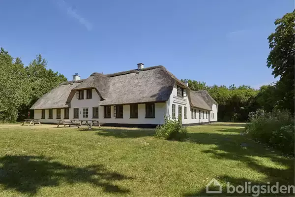 Stort hvidt bindingsværkshus med stråtag står i en grøn have omgivet af træer. Flere bænke står foran huset under en blå himmel.