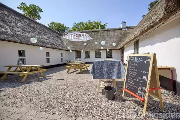 En gårdhave med stråtag, hvor tre picnicborde og en paraply står. En tavle menu viser "DRINKS: Rabarber, Hvidvin med rabarber, Ingefær, Citron med mynte". Lyskugler hænger ovenover.